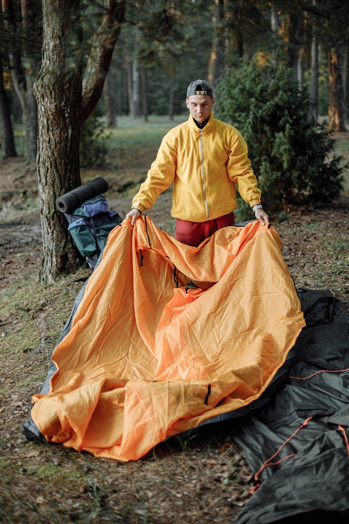 A young man sets up a bright orange tent in a peaceful forest campsite, preparing for an adventure.