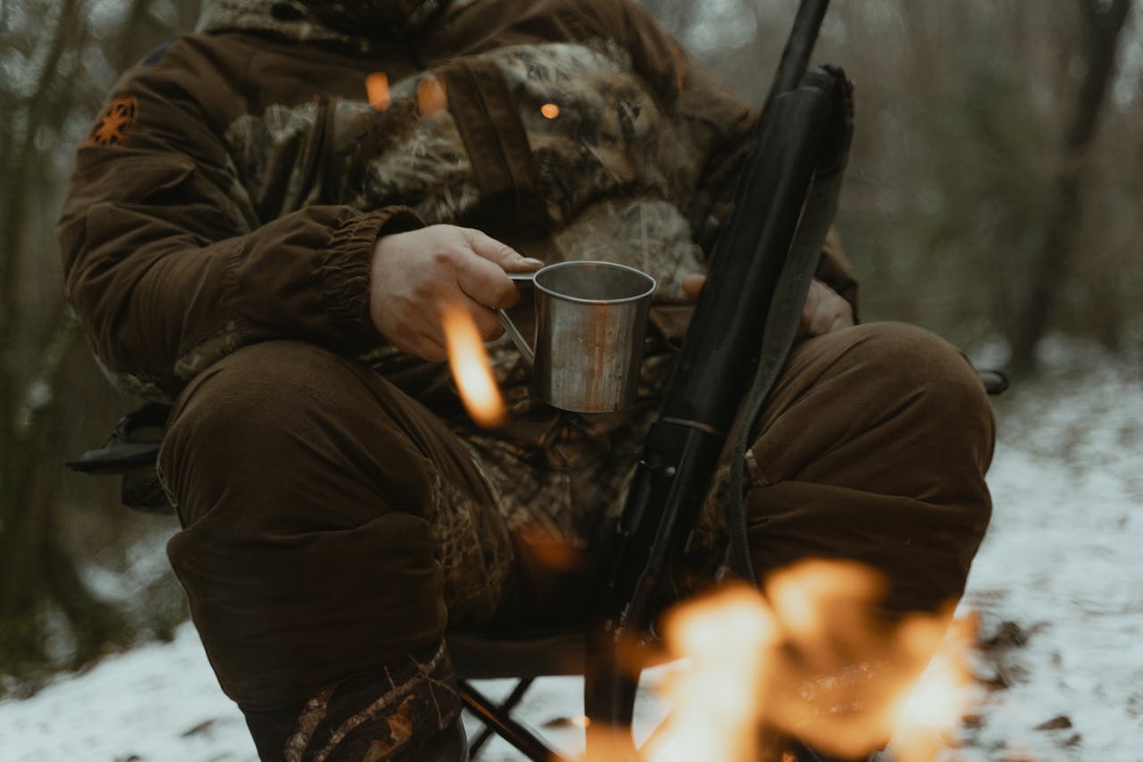 A hunter wearing camouflage sits by a campfire holding a metal mug in a snowy forest.