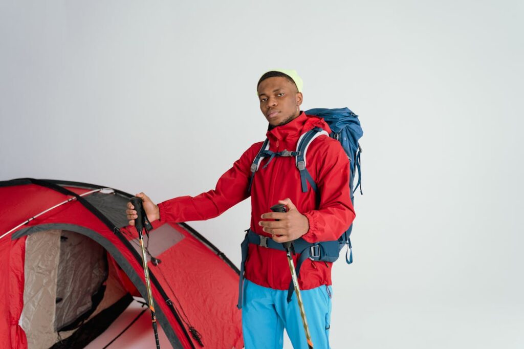 Male hiker in red jacket with backpack and ski poles beside a tent, ready for adventure.